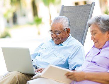 Two older persons reclining on lounge chairs; man looking at laptop, woman reading book.