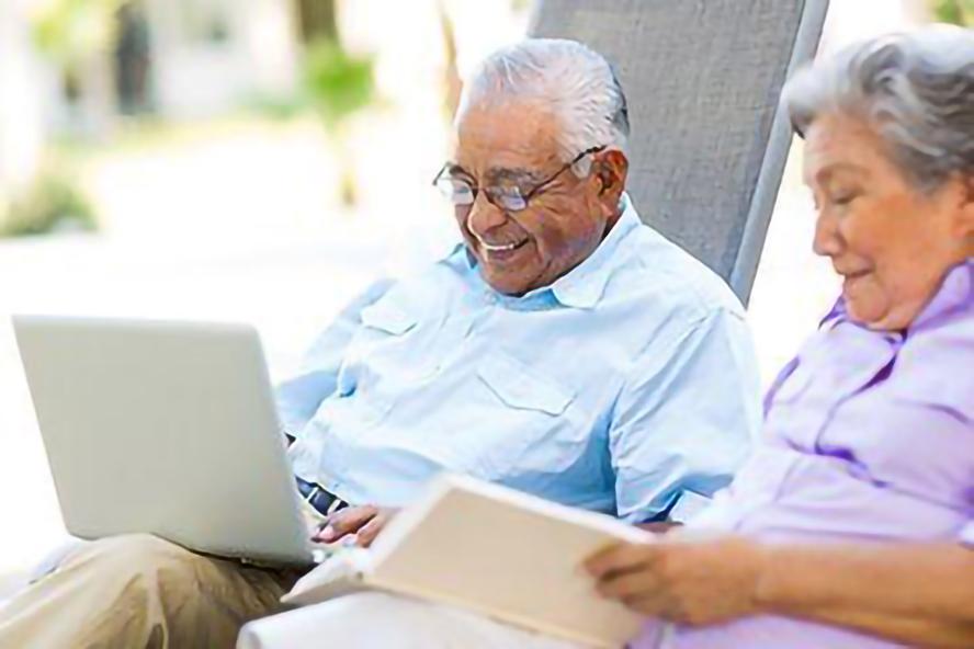 Two older persons reclining on lounge chairs; man looking at laptop, woman reading book.