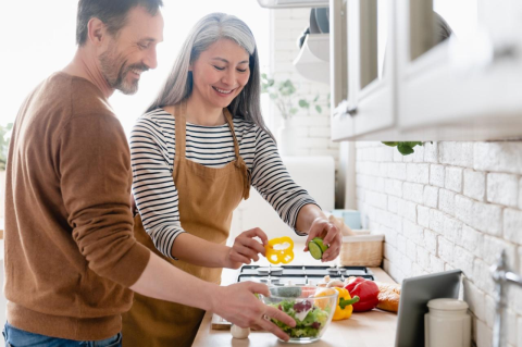 Older man in orange sweater and older woman with long white hair in orange apron and striped shirt  drop colorful vegetables into glass bowl in a white kitchen