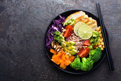 A black bowl filled with grilled chicken, brown rice, avocado, tomato, broccoli, red cabbage, chickpeas, and nuts.